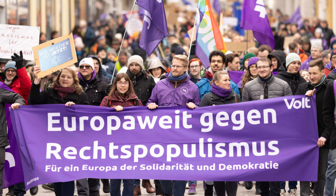 Demonstrationszug mit vielen Menschen, die Transparente und lila Fahnen der Bewegung Volt tragen. Im Vordergrund ein großes Banner mit der Aufschrift: „Europaweit gegen Rechtspopulismus – Für ein Europa der Solidarität und Demokratie.“ Weitere Schilder im Hintergrund, unter anderem mit den Botschaften „Nie wieder ist jetzt!“ und „Gegen Rassismus – für Menschlichkeit.“ Die Szene spielt auf einer Straße in einer Stadt.