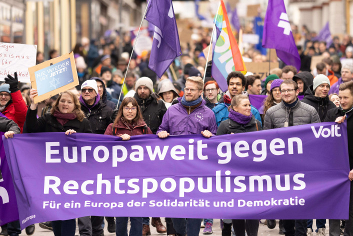 Demonstrationszug mit vielen Menschen, die Transparente und lila Fahnen der Bewegung Volt tragen. Im Vordergrund ein großes Banner mit der Aufschrift: „Europaweit gegen Rechtspopulismus – Für ein Europa der Solidarität und Demokratie.“ Weitere Schilder im Hintergrund, unter anderem mit den Botschaften „Nie wieder ist jetzt!“ und „Gegen Rassismus – für Menschlichkeit.“ Die Szene spielt auf einer Straße in einer Stadt.