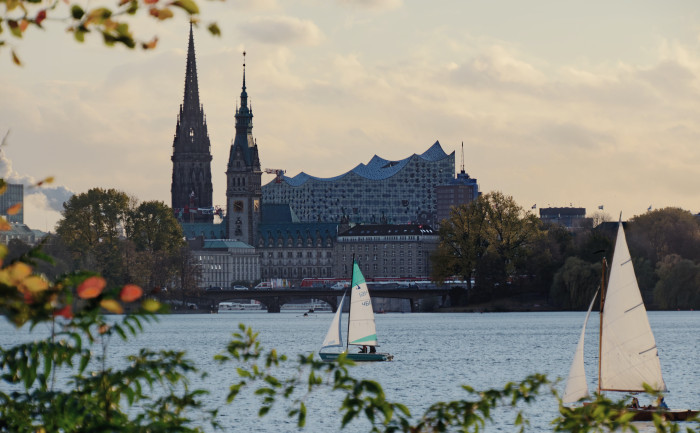 Der Blick über die Alster zeigt zwei Segelboote, eine Brücke, das Rathaus und im Hintergrund überragt die Elbphilharmonie.