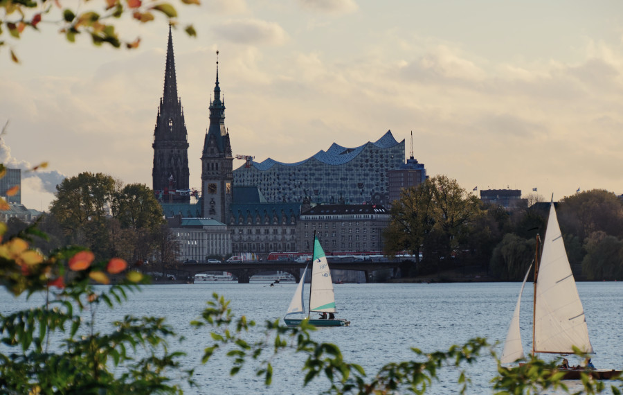 Der Blick über die Alster zeigt zwei Segelboote, eine Brücke, das Rathaus und im Hintergrund überragt die Elbphilharmonie.