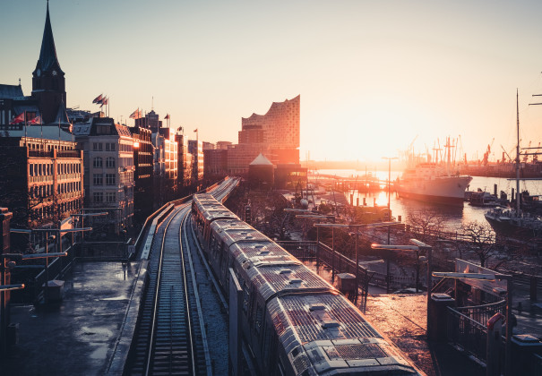 Eine Drohnenaufnahme hat Hamburg bei Sonnenaufgang eingefangen. Zur rechten Teile eine Schiff und Teile des Hafens, geradeaus die Elbphilharmonie und unter uns die Schienen der U-Bahn, sowie eine U-Bahn, die hier gerade durchrattert.